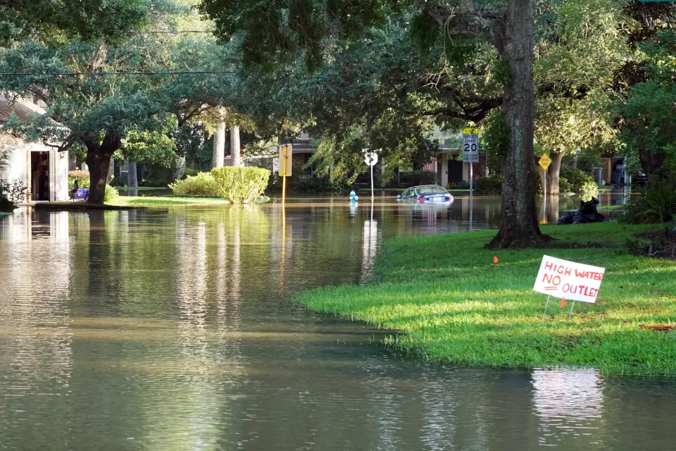 Flooded residential street with houses, yards, and trees on each side, a yard sign that reads "high water, no outlet," and a flooded car in the distance.